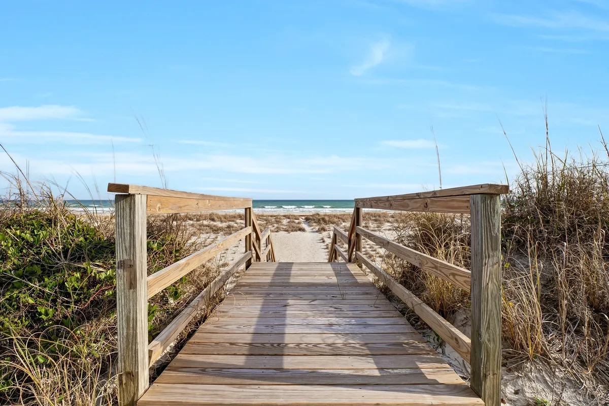 This image captures a wooden walkway leading directly to the ocean, with dune grass on the sides and the waves visible in the background. The scene evokes the anticipation and excitement of stepping onto the beach right from your accommodations.