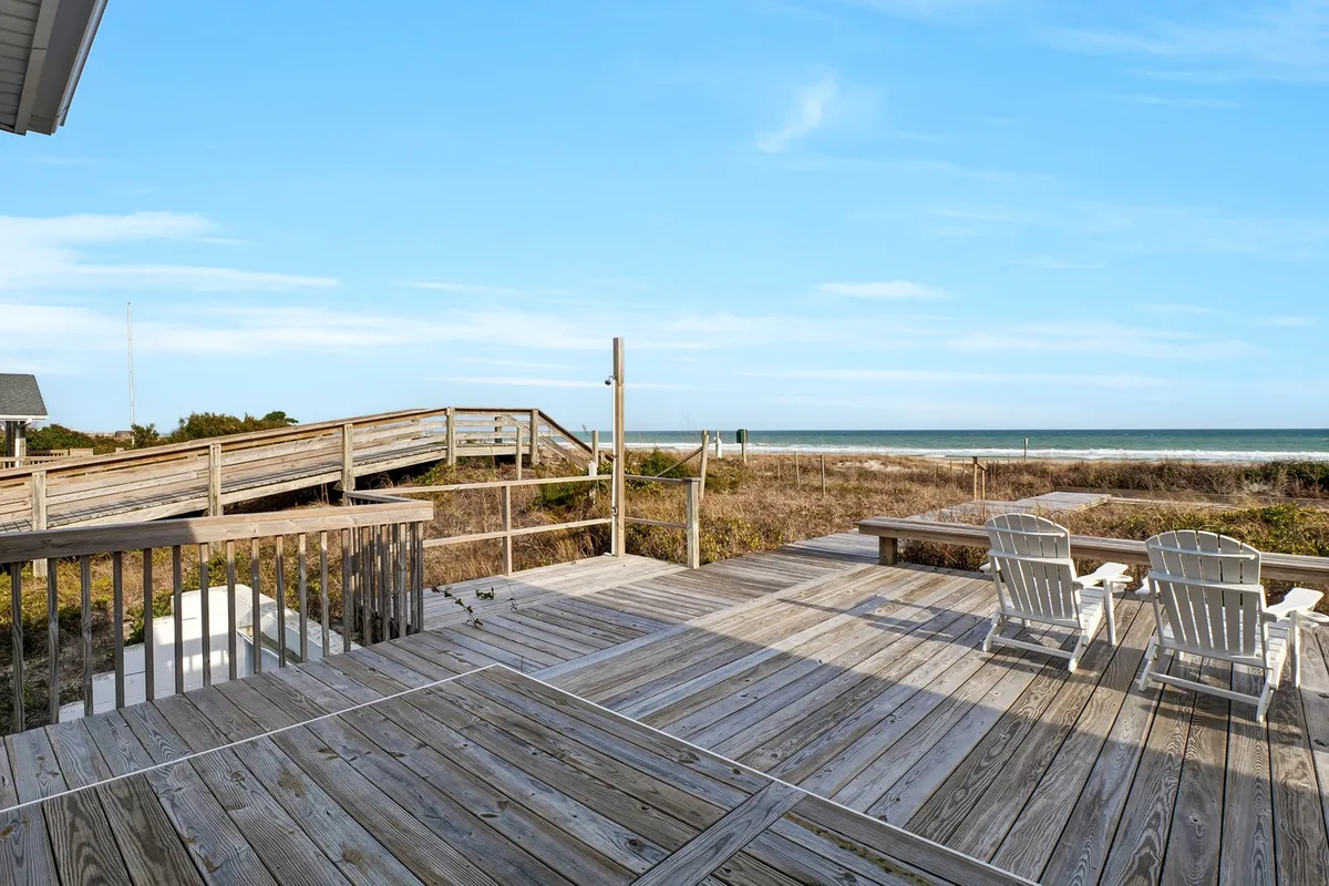 This image showcases a stunning beach view from a wooden deck furnished with adirondack chairs. The ocean is visible in the background, and the setup invites guests to relax, sunbathe, or watch the sunrise and sunset while enjoying the fresh sea breeze. The open deck directly connects the accommodation to the coastal environment, emphasizing the 'relaxation with a view' focus.