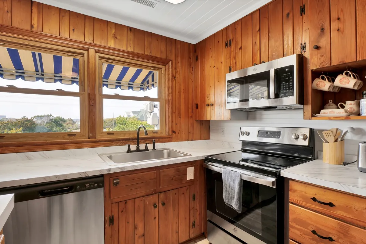 A sunlit kitchen featuring wooden cabinetry, modern stainless steel appliances, and an expansive marble countertop. Large windows with a striped awning flood the space with natural light. The layout encourages communal cooking and family gatherings, as bar seating invites guests to linger or help with meal preparation.