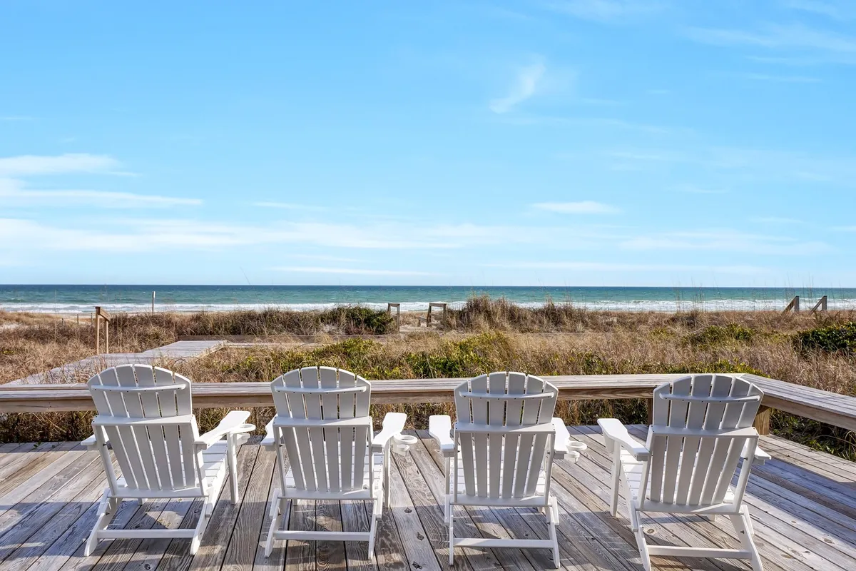 The image shows a tranquil exterior scene featuring classic white Adirondack chairs set atop a weathered wooden deck, overlooking sweeping ocean views framed by soft beach grass. The Atlantic glistens in the background beneath a pastel sunrise, offering a serene space perfect for relaxation and taking in the natural beauty of Topsail Beach.