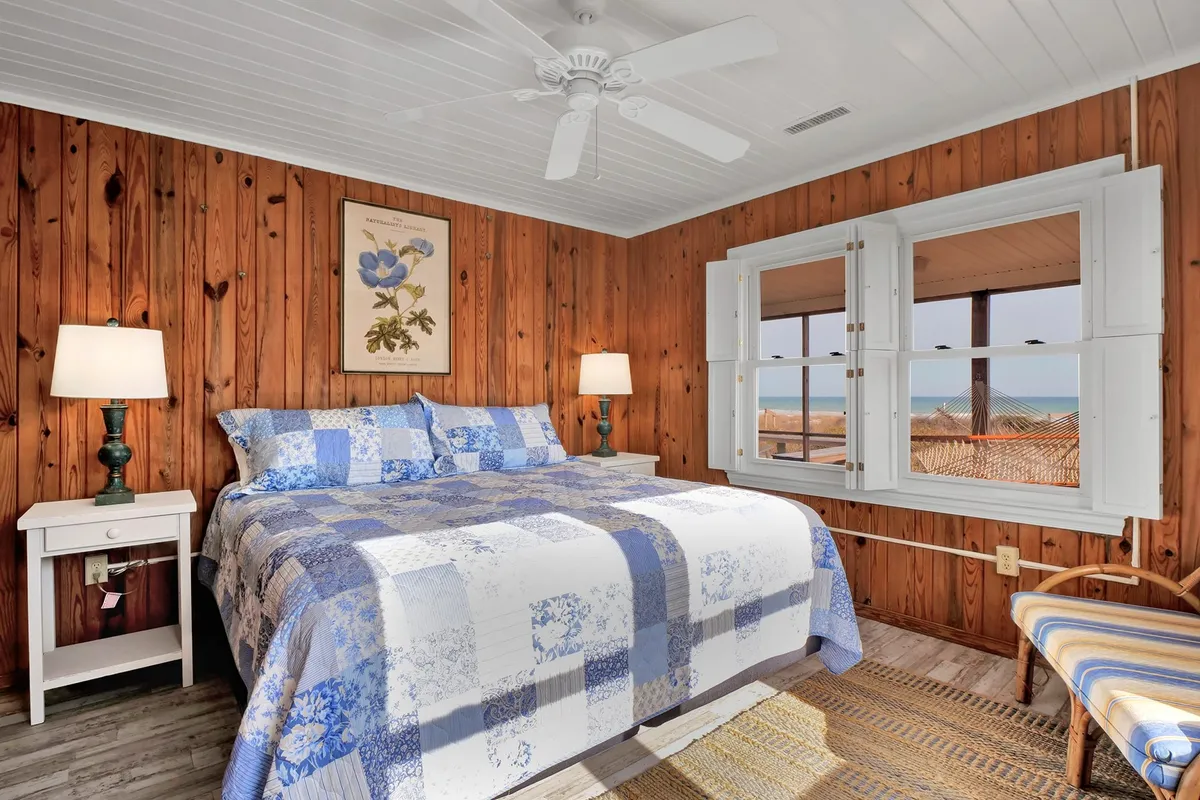 A bedroom featuring a bed with a blue and white patchwork quilt, two bedside lamps, framed botanical prints, and open windows revealing an inviting view. The ambiance is serene and comfortable, perfect for a peaceful night's rest.