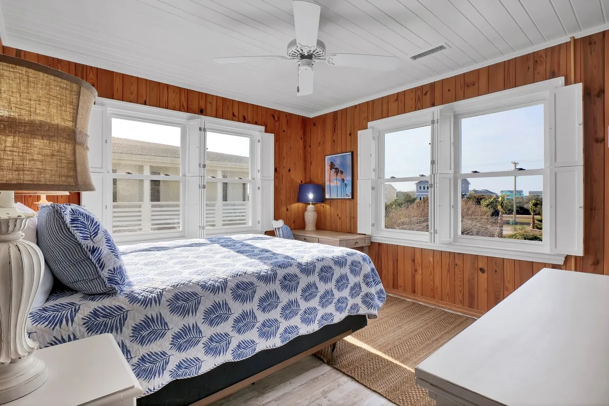 A tranquil bedroom with blue and white patterned bedding, large windows streaming in natural light, wooden wall paneling, and a warm bedside lamp. The space feels airy and peaceful, perfect for relaxing after a day at the beach.