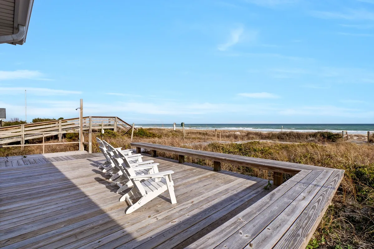 A sunlit wooden deck outfitted with white Adirondack chairs, perfectly angled to maximize the sweeping ocean views. The inviting seating arrangement and clear sightlines to the water evoke a sense of calm and connection to the beach.