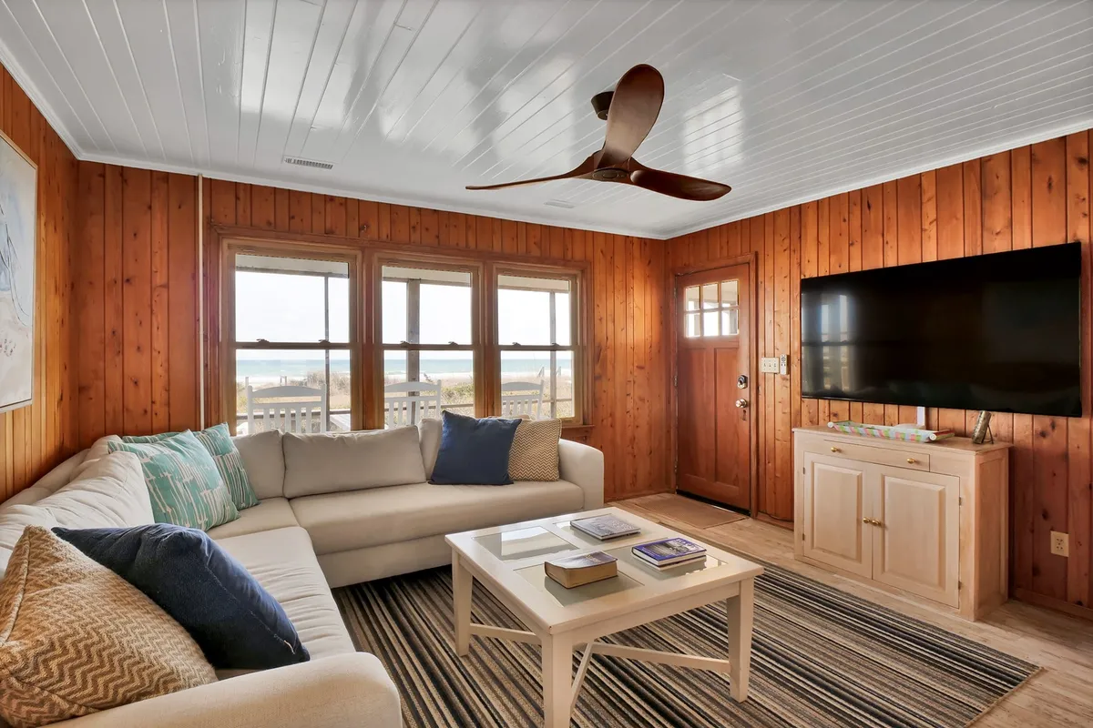 The image features a comfortable sectional sofa set near large windows, with a flat-screen TV and a coffee table topped with books. Natural light floods the room, and the décor is warm and inviting, emphasizing a relaxed and homely atmosphere—perfect for movie nights or unwinding after beach adventures.
