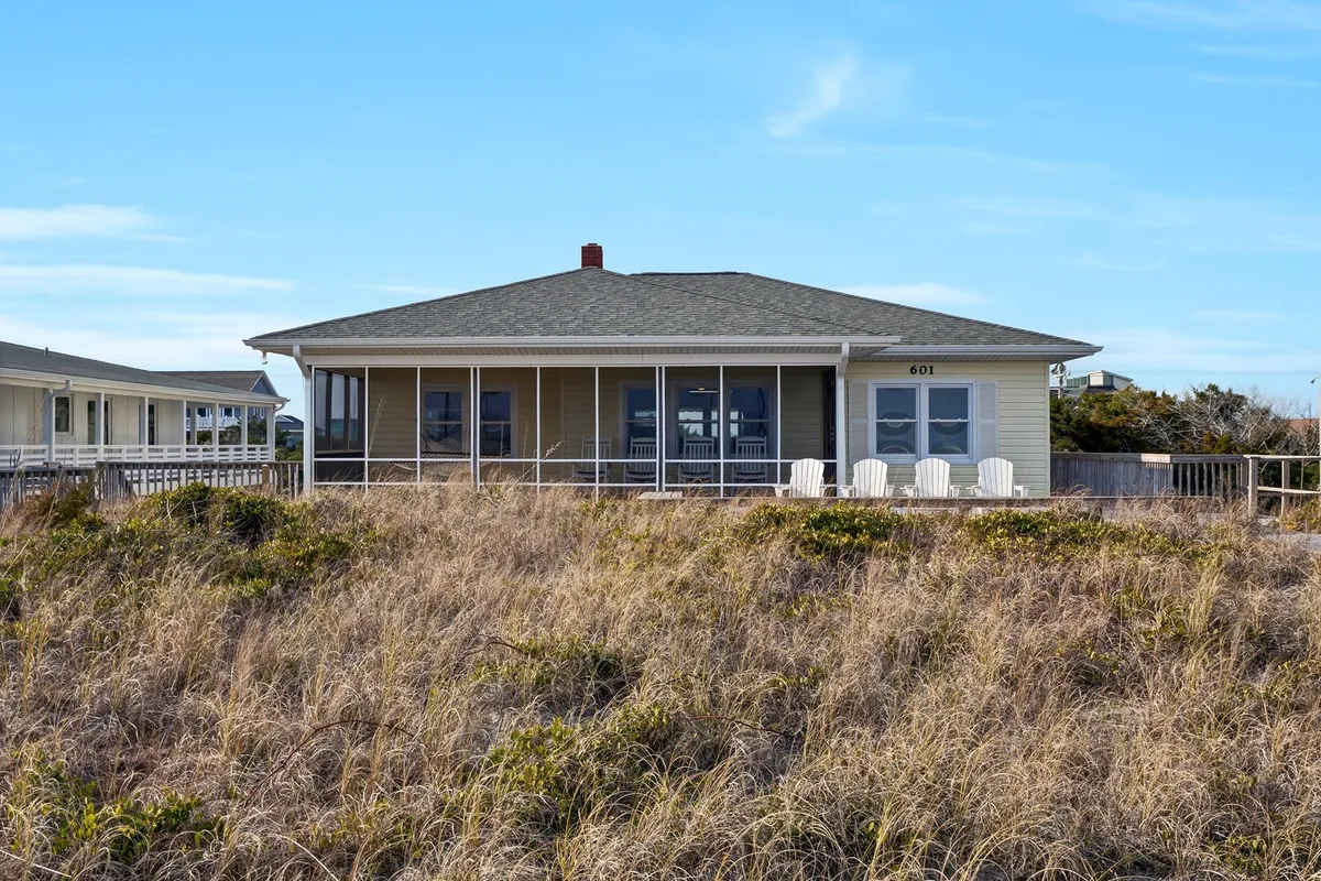 The photo features the house’s exterior, with a clear view of the welcoming front porch, chairs arranged for conversation, and the surrounding landscape. The porch is designed for gatherings, with plenty of space for guests to relax and enjoy the coastal ambiance together.