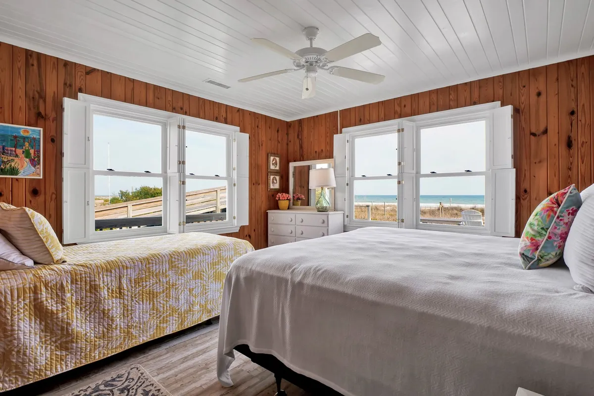 A cozy bedroom with two beds positioned beside large windows that offer an ocean view. The wooden wall panels, decorative lamp, and framed artwork create a warm and inviting setting ideal for families or groups.
