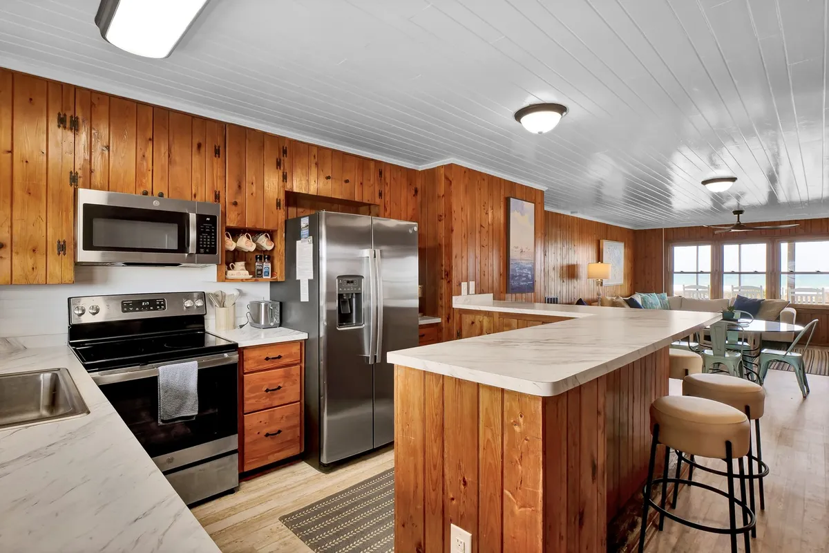 A cozy kitchen interior with a central island, wood paneling, and large windows that let in natural light. Stainless steel appliances are integrated into the cabinetry, offering functional cooking space. The setting is ideal for socializing and meal prep, with room for gathering and sharing stories.