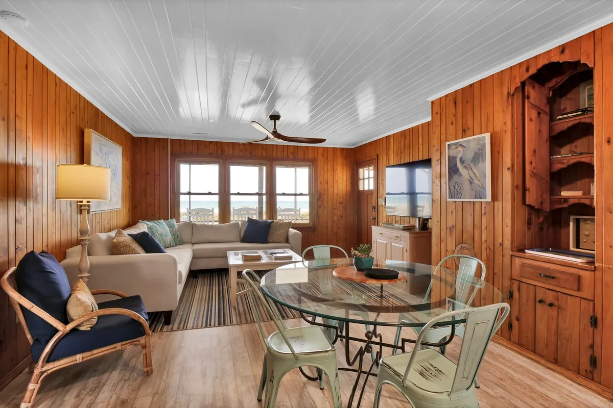 This interior image showcases a spacious open-plan living and dining area, featuring a large sectional sofa, a glass dining table, wood-paneled walls, and expansive windows that let in abundant natural light. The layout invites gatherings and relaxation, with clear sightlines between the living and dining spaces, creating a seamless flow ideal for socializing.