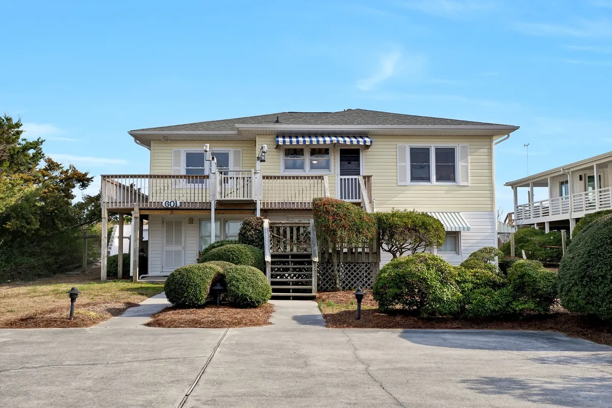This image displays a welcoming front porch with a classic railing, well-kept bushes, entrance steps, and awnings over the windows. The porch offers space for seating and relaxation, while the manicured garden and the structure’s proximity to the outdoors evoke a direct connection to nature and the beach environment.