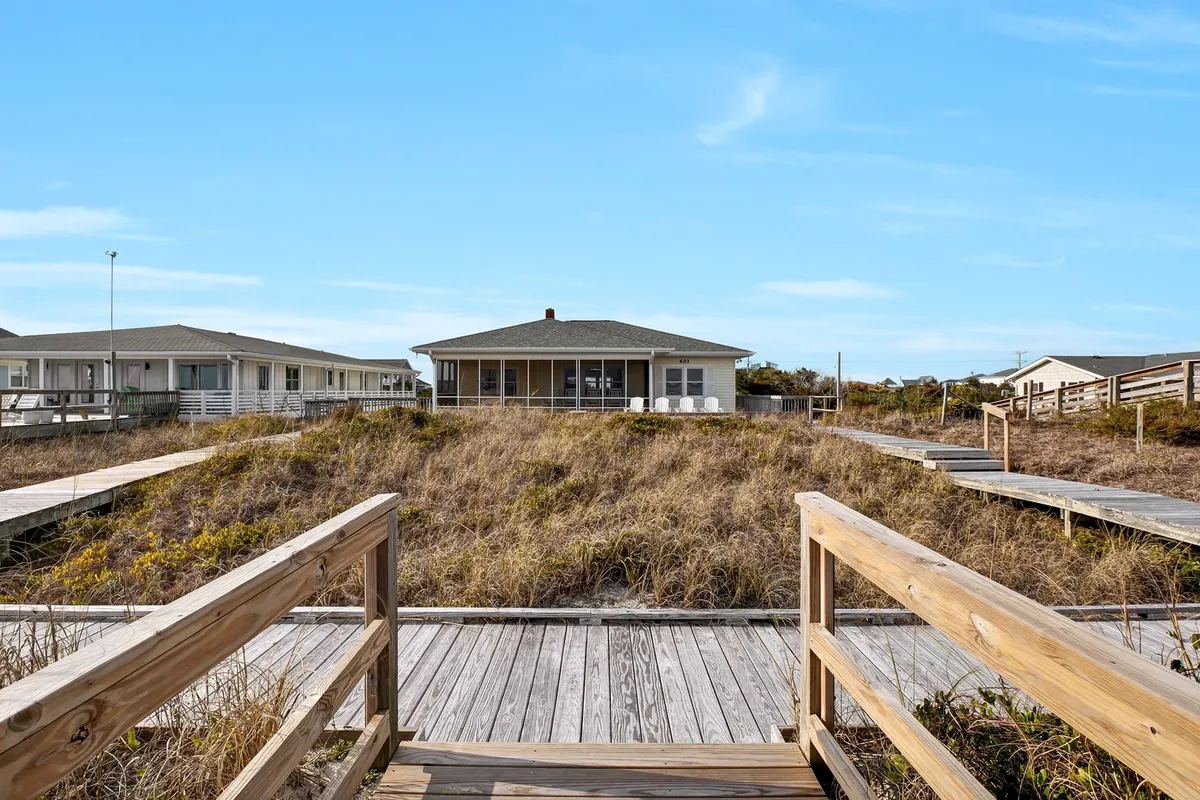A view of a wooden walkway surrounded by grass leading toward the house structure, evoking the sense of returning from a beach adventure. The scene suggests relaxation after outdoor activities, with space for family gatherings or picnics.